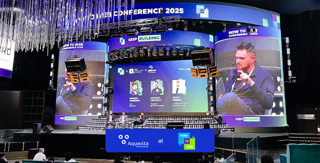 Conference stage at How To Web 2025 with a panel discussion on screen and the Aquavita banner