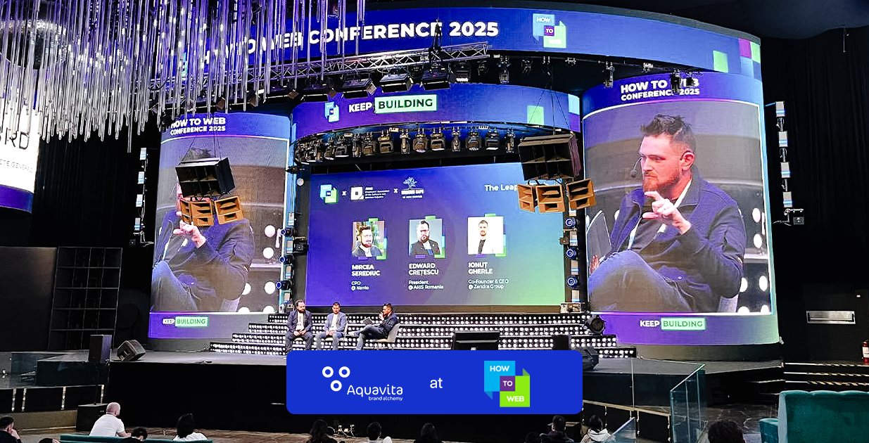 Conference stage at How To Web 2025 with a panel discussion on screen and the Aquavita banner